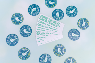 Die-cut Six Byrd Cider Company stickers scattered around printed sticker sheets